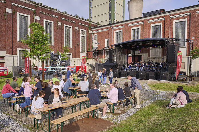 Kauenhof bei Tag mit aufgebauter Bühne und Menschen, die auf Bierzeltgarnituren sitzen.