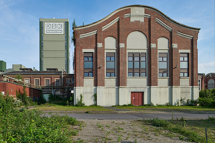 Frontalansicht der Zentralmaschinenhalle, ein rotes Backsteingebäude, mit einer Straße und einer Grünfläche davor.