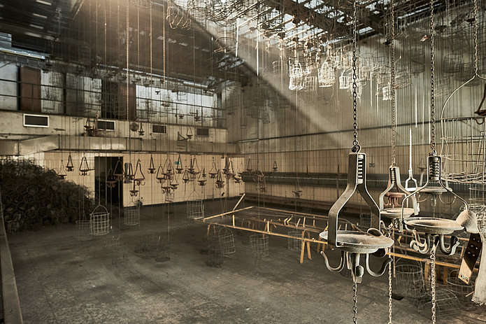 Innenraum der Kaue mit von der Decke hängenden Ketten und Kauenkörben. Diffuses Licht fällt in den Raum.