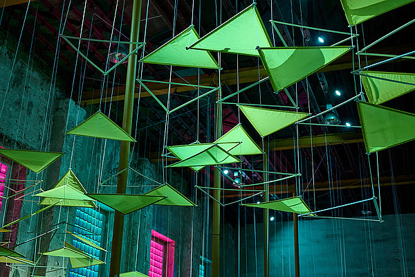 Installation aus neonfarbenen, leuchtenden Dreiecken an der Decke eines dunklen Raums.
