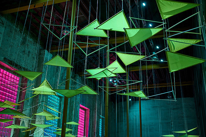 Installation aus neonfarbenen, leuchtenden Dreiecken an der Decke eines dunklen Raums.