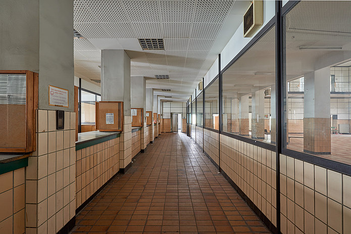 Innenansicht der Lohn- und Lichthalle: langer Flur mit braunen Fliesen an Boden und Wand, links mehrere Durchreichen zum Nebenraum, rechts verglasten Wänden zum Nebenraum.