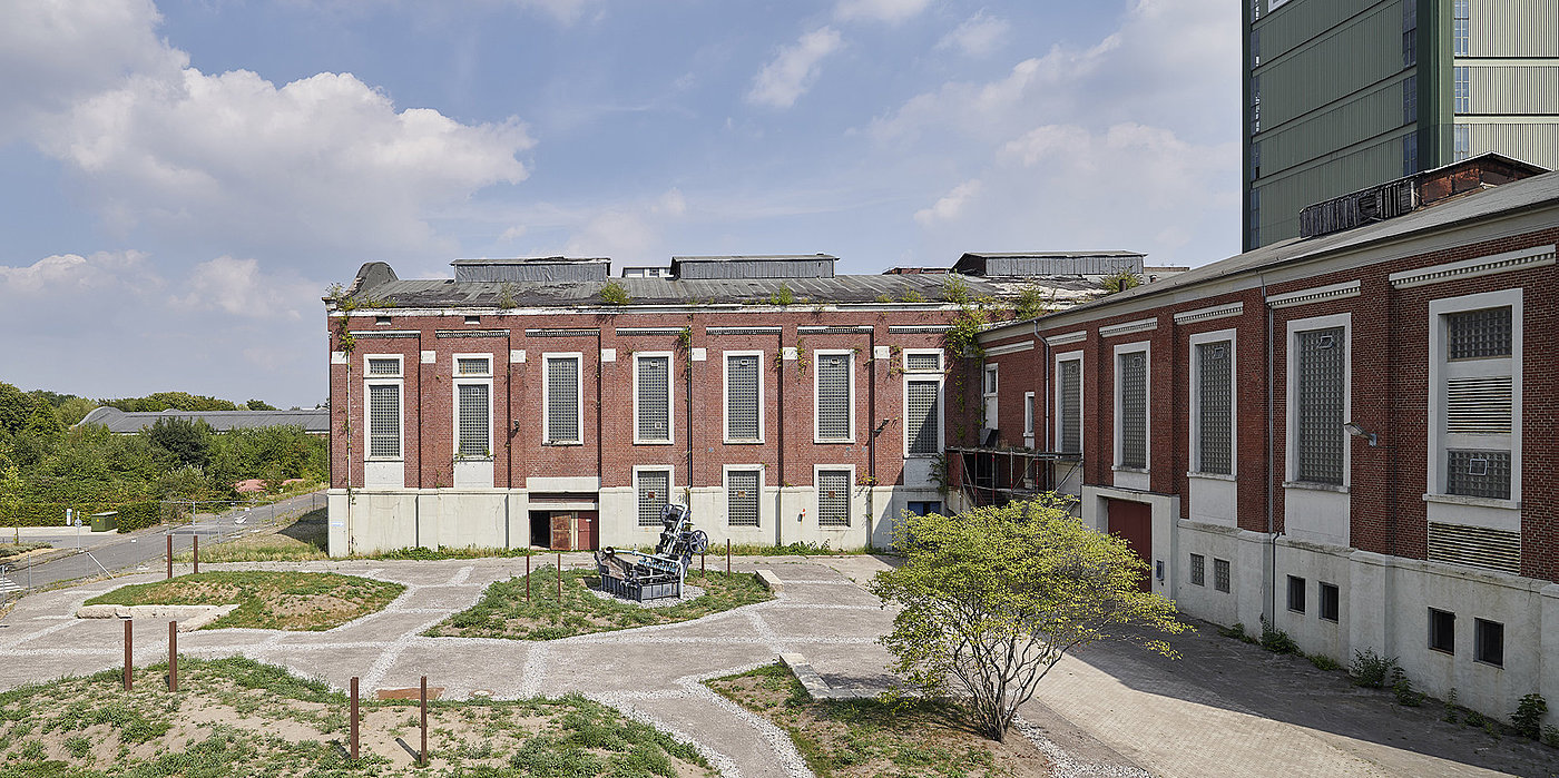 Blick auf den Kauenhof mit partiellen Grünflächen und einem Baum. Vor einem roten Backsteingebäude steht die Installation „Kaskaden Wasserspiel“.