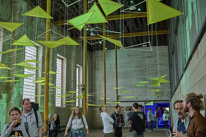 Deckeninstallation aus neonfarbenen Dreiecken in einem hellen Raum; Besucher gehen darunter entlang, unterhalten sich und betrachten die Installation.