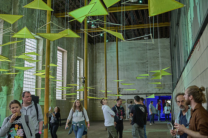 Deckeninstallation aus neonfarbenen Dreiecken in einem hellen Raum; Besucher gehen darunter entlang, unterhalten sich und betrachten die Installation.