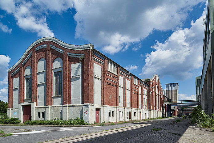 Seitliche Ansicht der Zentralmaschinenhalle mit sichtbaren Leitungen, die zu Nebengebäuden führen; dazwischen verläuft eine Straße.