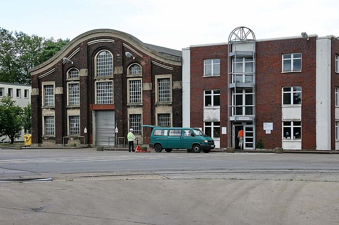 Hochregallager von außen, zwei rote Backsteinhäuser, im Vordergrund ein türkiser Bulli und ein Mann mit Helm und Warnweste