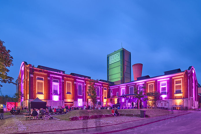 Kauenhof bei Nacht: Rotes Backsteingebäude leuchtet in Neon-Orange und Pink. Vor dem Gebäude ist eine Bühne aufgebaut, Menschen sitzen auf Bierzeltgarnituren.