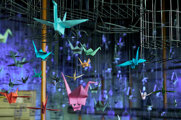 Neonfarbene Origami-Kraniche leuchten als Lichtinstallation, aufgehängt an Kauenkörben in der Schwarzkaue