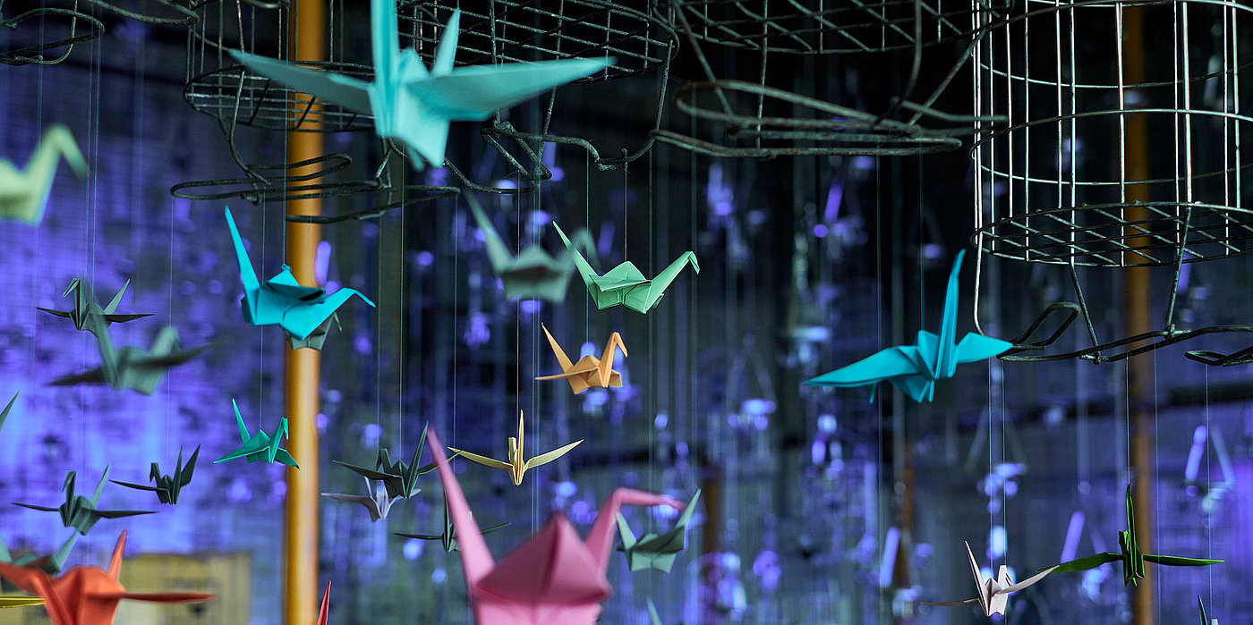 Neonfarbene Origami-Kraniche leuchten als Lichtinstallation, aufgehängt an Kauenkörben in der Schwarzkaue