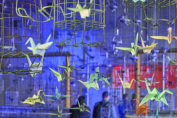 Leuchtende Origami-Kraniche in Neonfarben hängen an Kauenkörben in der Schwarzkaue; im Hintergrund sind unscharf Silhouetten von Menschen zu sehen.