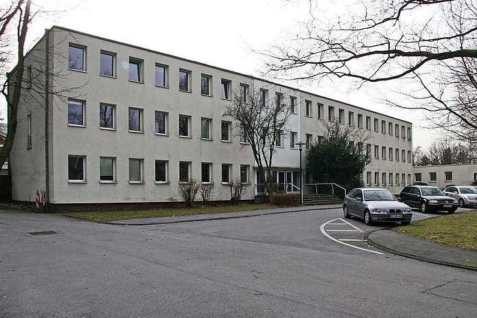 Verwaltungsgebäude Außenansicht, längliches weißes Gebäude mit geparkten Autos davor