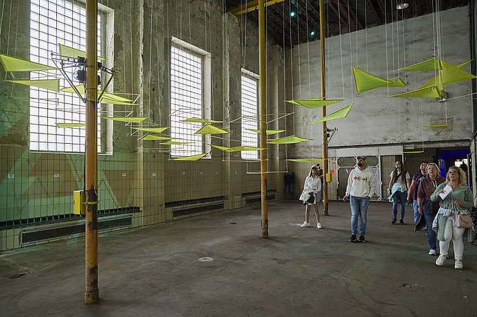 Deckeninstallation aus neonfarbenen Dreiecken in einem hellen Raum; Besucher gehen darunter entlang.