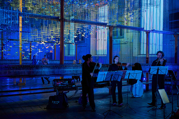 In der dunklen Schwarzkaue spielen drei Geiger:innen unter hell leuchtenden, neonfarbenen Origami-Kranichen, die an Kauenkörben hängen.