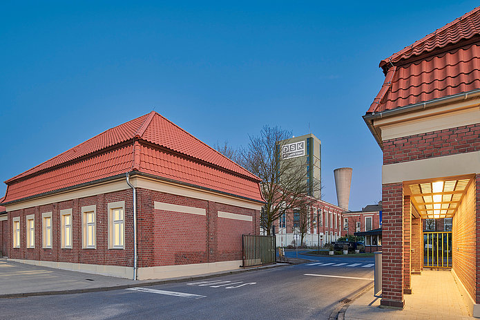 Abendliche Ansicht des geöffneten Tores zwischen den Torhäusern. Warme gelbe Lichter sind eingeschaltet und beleuchten die Szene.