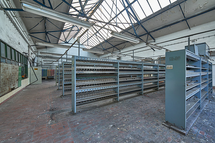 Innenansicht der Lohn- und Lichthalle mit einer Reihe von Regalen. An der Decke befinden sich Fenster und lange Hängeleuchten.