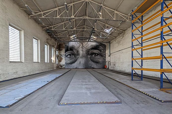 Hochregallager von innen, eine helle Halle mit rechts gelben Regalen und am Ende einer großen schwarz-weiß Nahaufnahme eines Gesichts