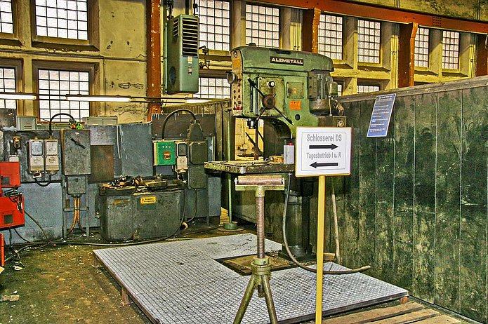 Kesselhaus 1 Innenansicht, grünes Regal, zwei verschiedene Maschinen und ein Schild, dass auf die Schlosserei und den Tagesbetrieb verweist.