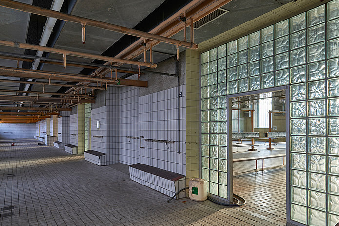 Flur vor der Kaue mit weißen Wandfliesen, Kupferrohren an der Decke und einem Durchgang mit quadratischen Glasbausteinen.